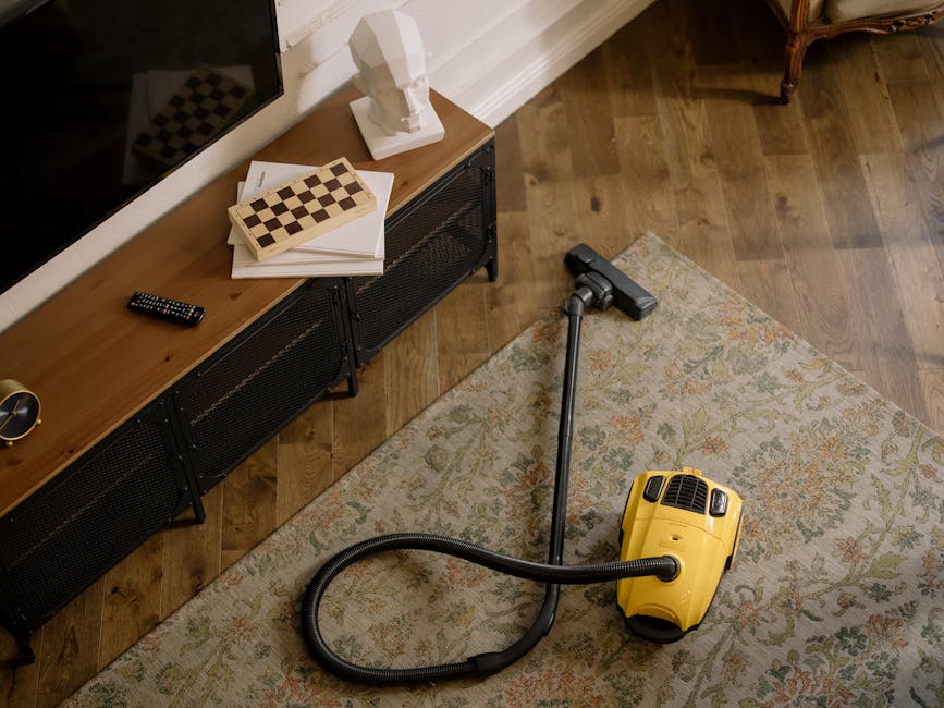 A yellow canister vacuum cleaner with a flexible black hose is positioned on a patterned area rug, which covers a wooden floor in a residential living room. The vacuum's hose extends toward a modern black metal and wooden TV stand, on top of which are a stack of white papers, a closed chessboard, a decorative white bust sculpture, and a black remote control. The room is well-lit, with natural light reflecting off the polished wooden floor and the clean, dust-free surfaces, exemplifying professional surface and deep cleaning practices by Carpet Cleaners W3 as part of their domestic cleaning services focused on maintaining hygiene and sanitation in Chelsea period homes.