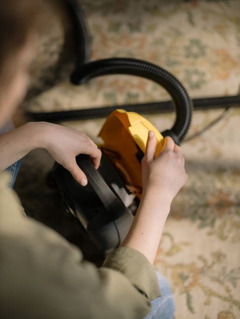 A person operating a yellow portable vacuum cleaner on a carpeted floor in a living room or similar interior space. The carpet features a detailed floral and foliage pattern in muted tones. The individual holds the vacuum's handle with one hand while adjusting the device with the other, focusing on surface cleaning to remove dust and debris. The background includes a section of a wooden floor and some indistinct furniture or decor, with soft, natural lighting illuminating the scene. The image emphasizes thorough domestic carpet cleaning as part of a detailed surface cleaning process, consistent with services offered by Carpet Cleaners W3 for Kings Road period homes in Chelsea, W3.