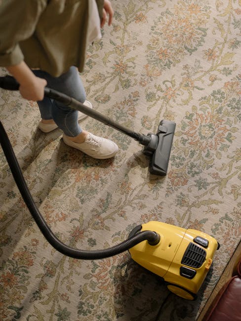 A yellow canister vacuum cleaner with a flexible black hose is positioned on a patterned area rug, which covers a wooden floor in a residential living room. The vacuum's hose extends toward a modern black metal and wooden TV stand, on top of which are a stack of white papers, a closed chessboard, a decorative white bust sculpture, and a black remote control. The room is well-lit, with natural light reflecting off the polished wooden floor and the clean, dust-free surfaces, exemplifying professional surface and deep cleaning practices by Carpet Cleaners W3 as part of their domestic cleaning services focused on maintaining hygiene and sanitation in Chelsea period homes.